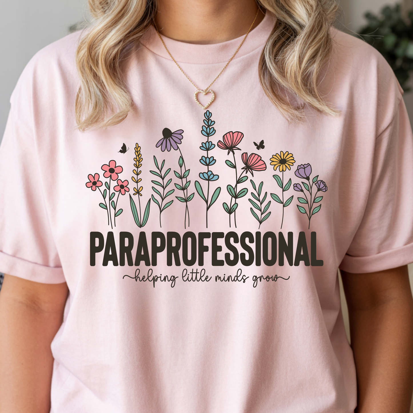A woman wearing a light pink short-sleeve t-shirt with a graphic of colorful wildflowers and the text 'PARaprofessional helping little minds grow'.