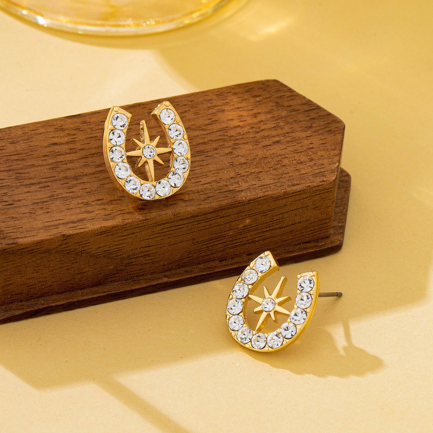 Gold earrings with star and horseshoe design on a wooden stand against a beige background