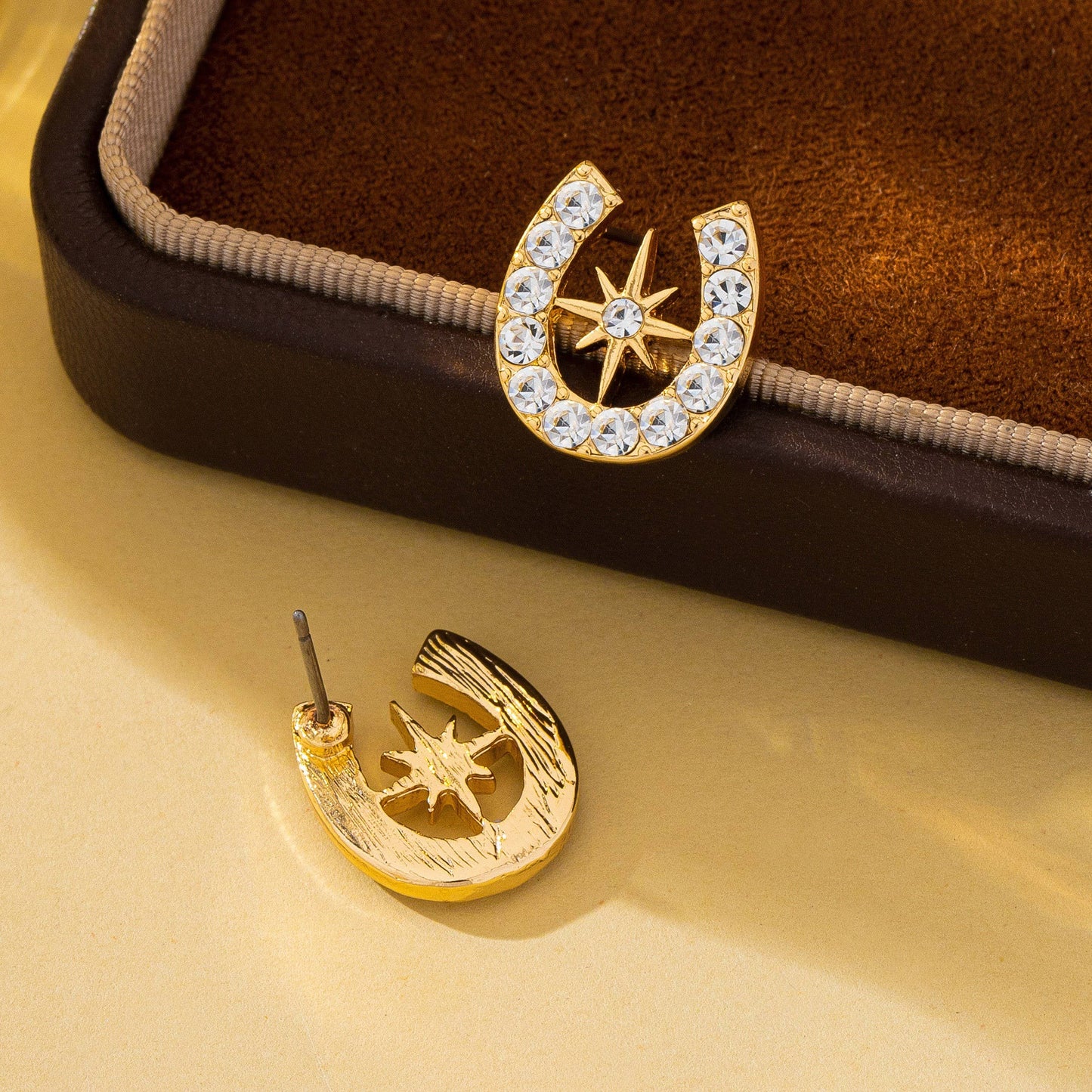Gold horseshoe and star earrings on a brown cushioned jewelry box.