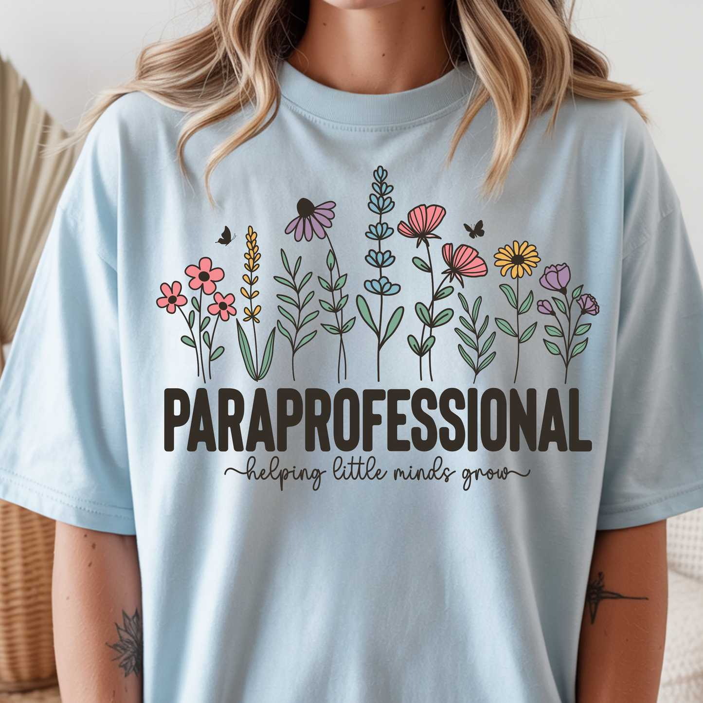 A woman wearing a light blue graphic T-shirt with a colorful line-art floral design and the text 'PARaprofessional helping little minds grow' on the front.