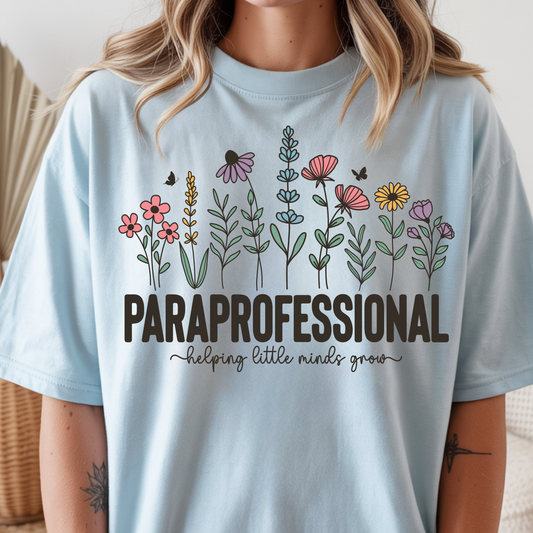 A woman wearing a light blue graphic T-shirt with a colorful line-art floral design and the text 'PARaprofessional helping little minds grow' on the front.